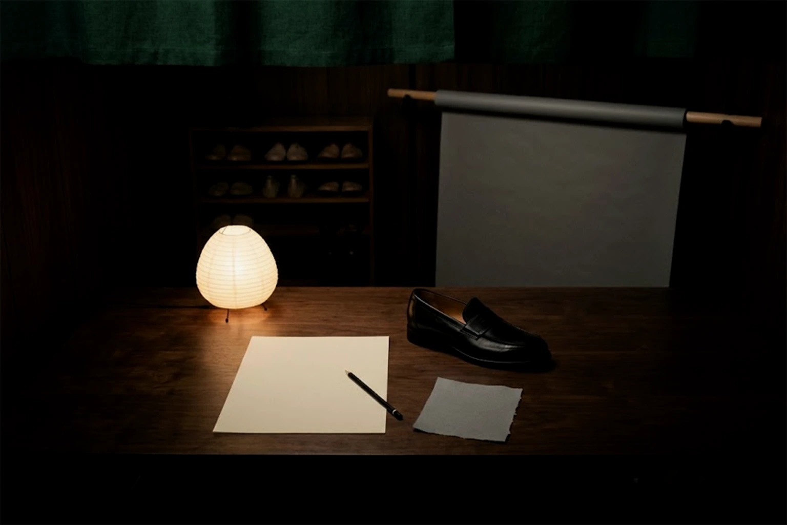 A sheet of blank paper and a pencil on a wooden desk under a paper lamp, beside a scrap of grey paper and one black loafer
