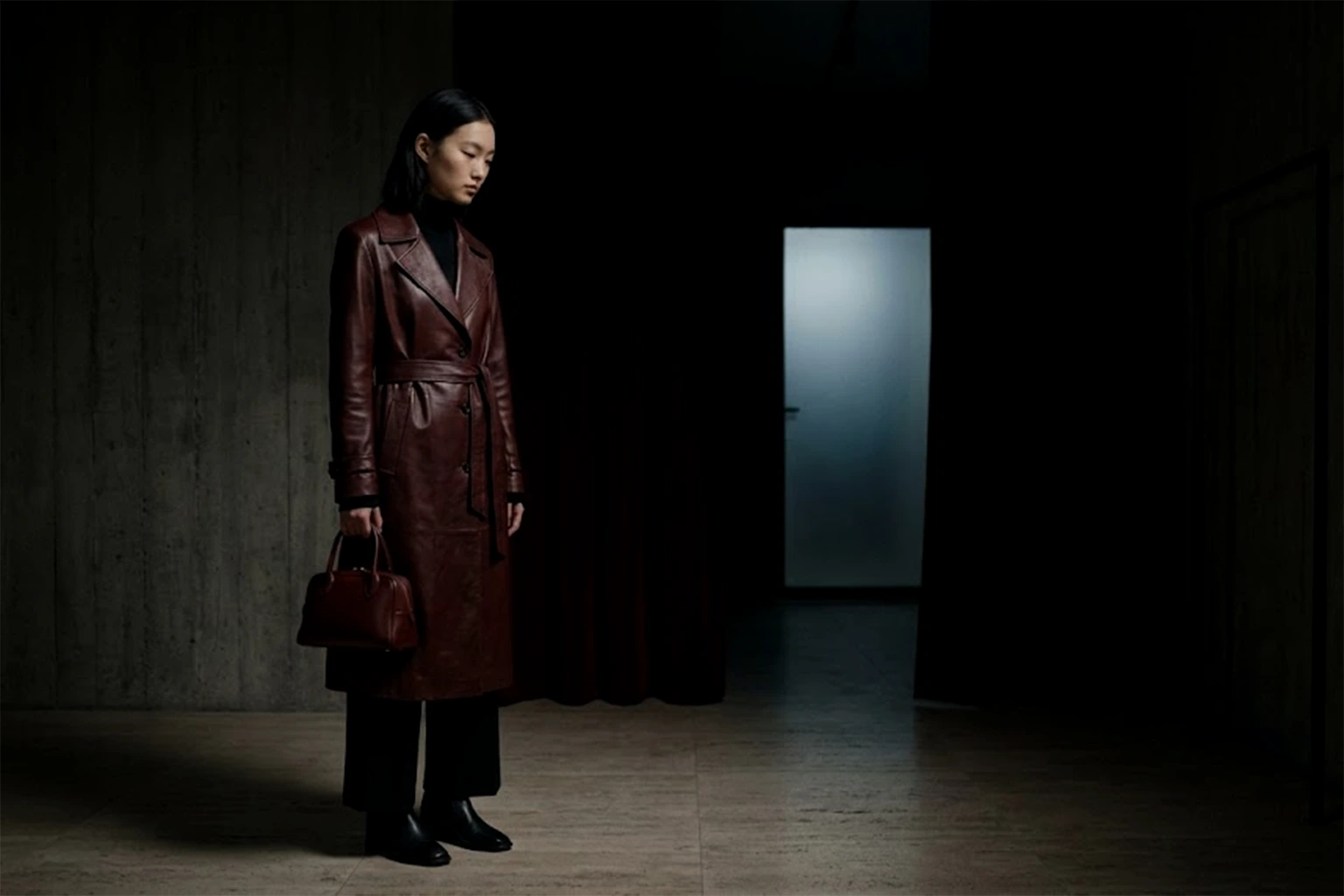In the dark back room, a woman in an oxblood leather trench coat stands alone, holding a small bag of the same colour