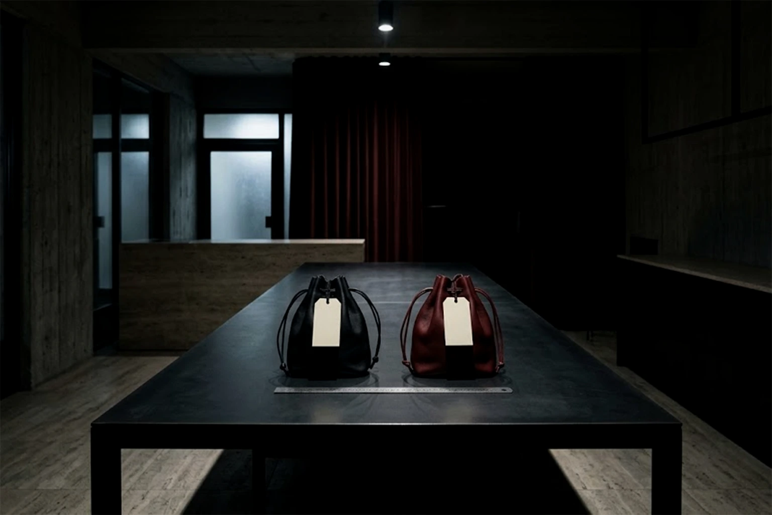 Two leather drawstring bags of the same shape, one black and one oxblood, side by side on a blackened-steel worktable with blank cream tags
