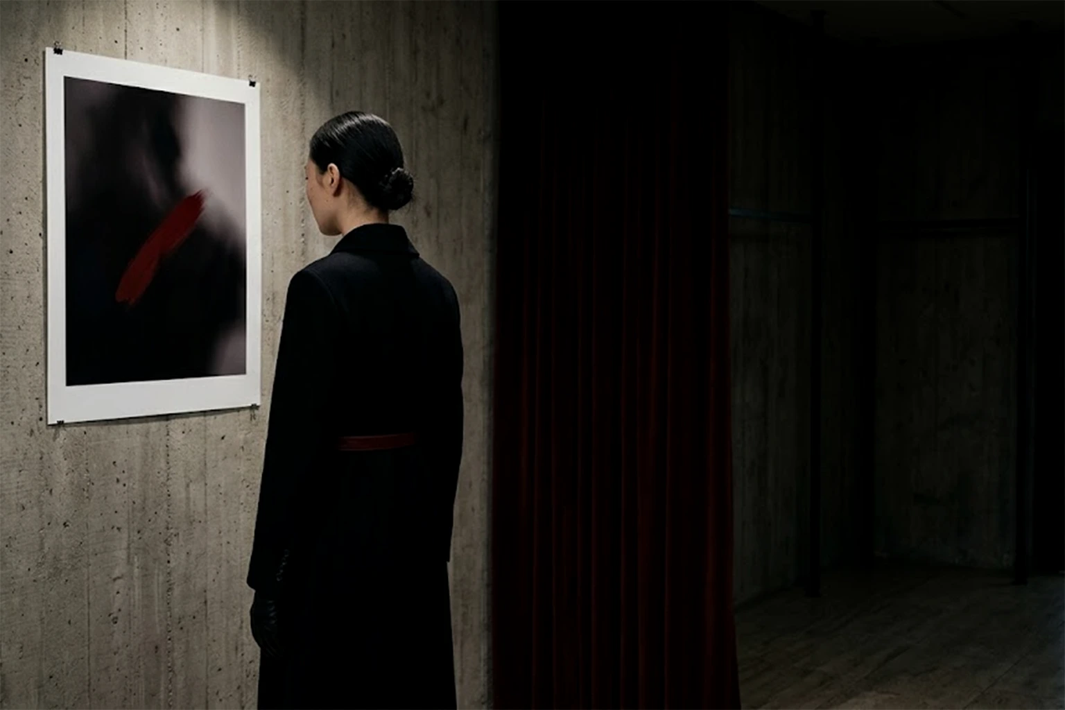 A woman in a black coat, seen from behind, looks at a single large print pinned to a concrete wall: a dark blur with one stroke of red